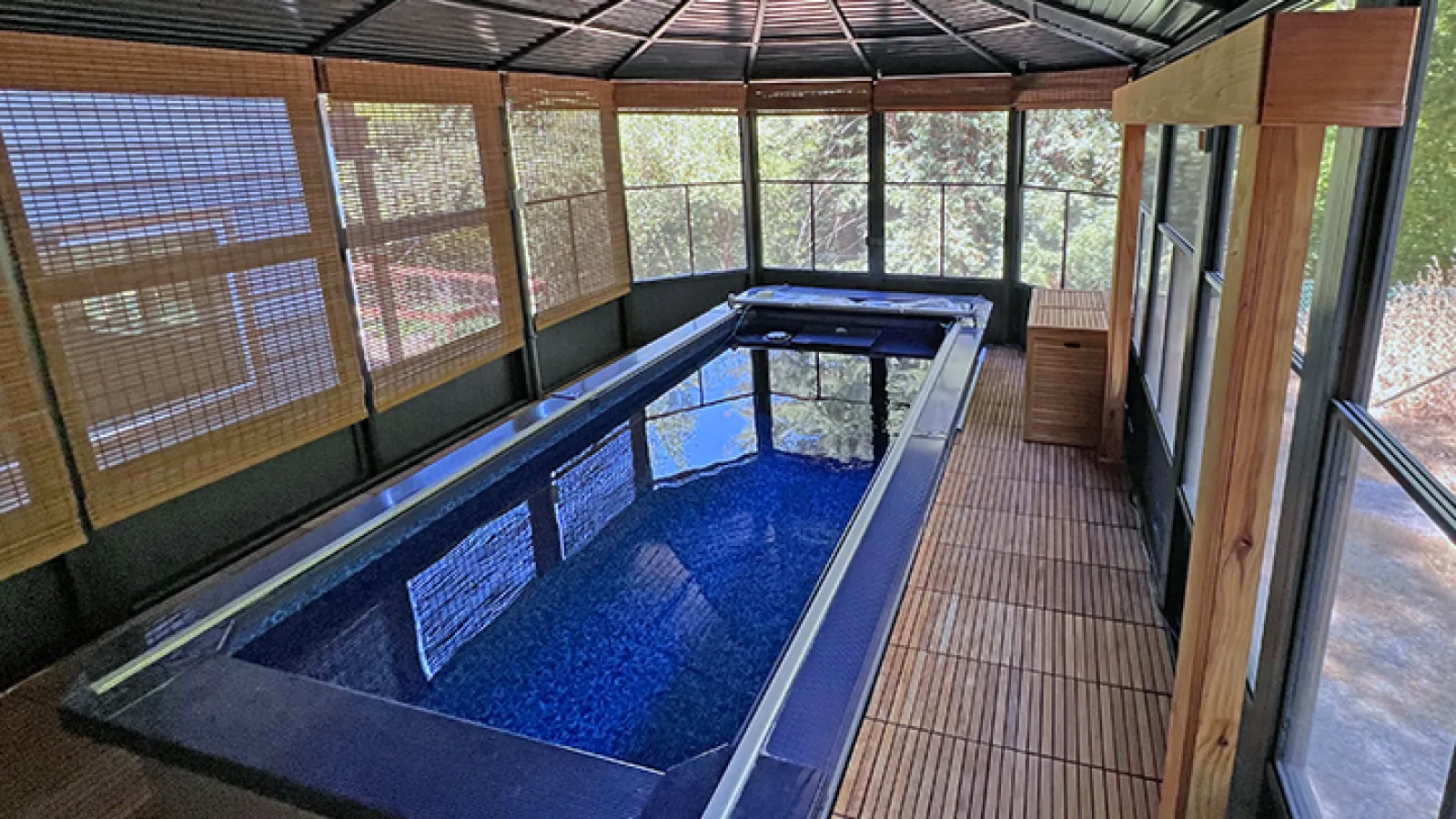 Indoor rectangular pool with blue water under a metal roof and surrounded by bamboo blinds and wooden flooring.