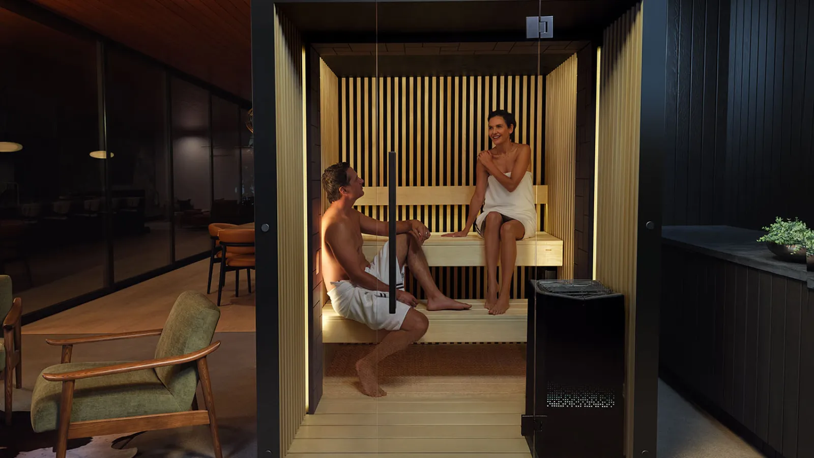 Two people relaxing inside a modern steam sauna with wooden interior and glass door in a home setting.