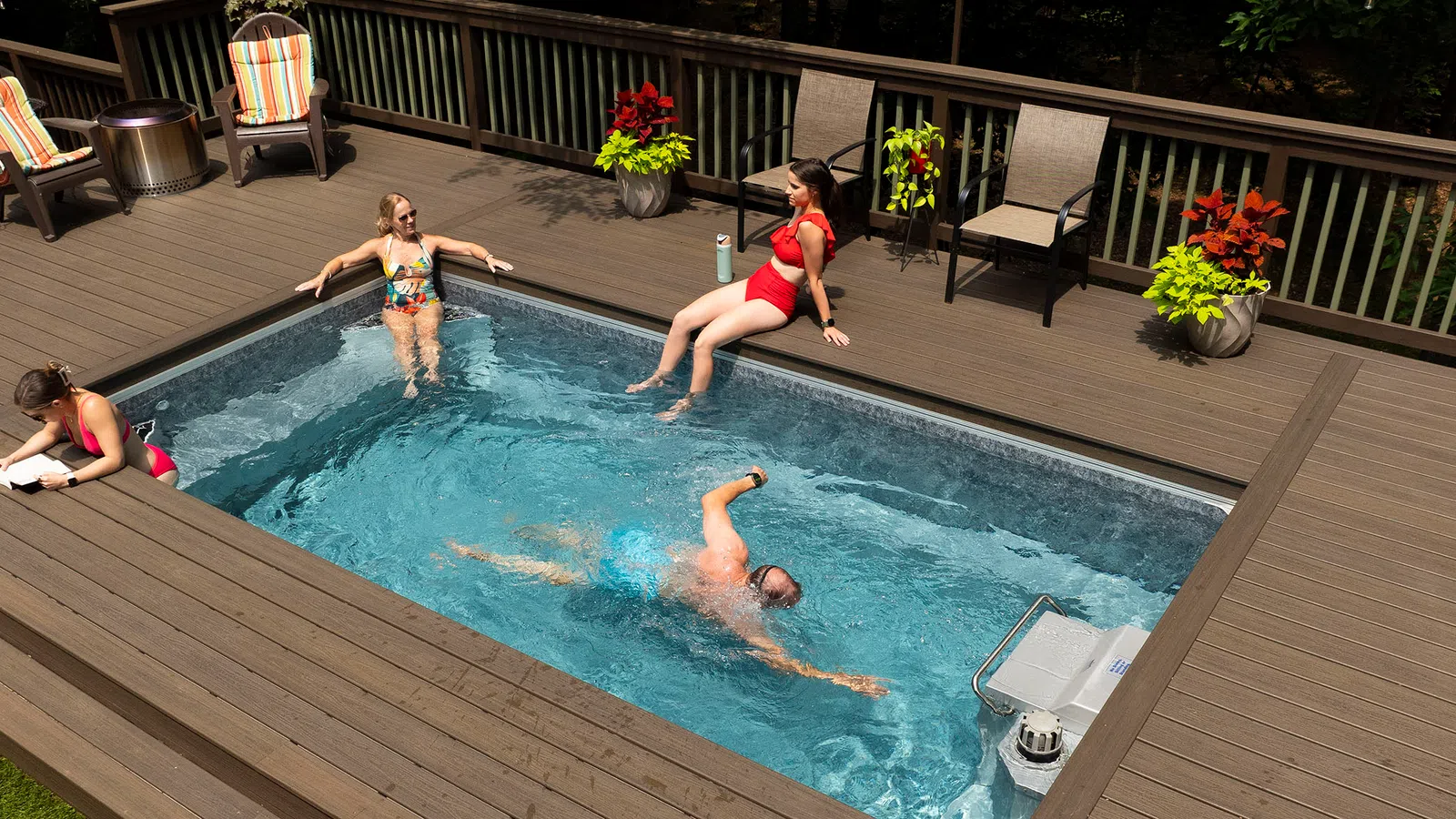 Four people enjoying a built-in backyard swimming pool with wooden deck and outdoor seating.