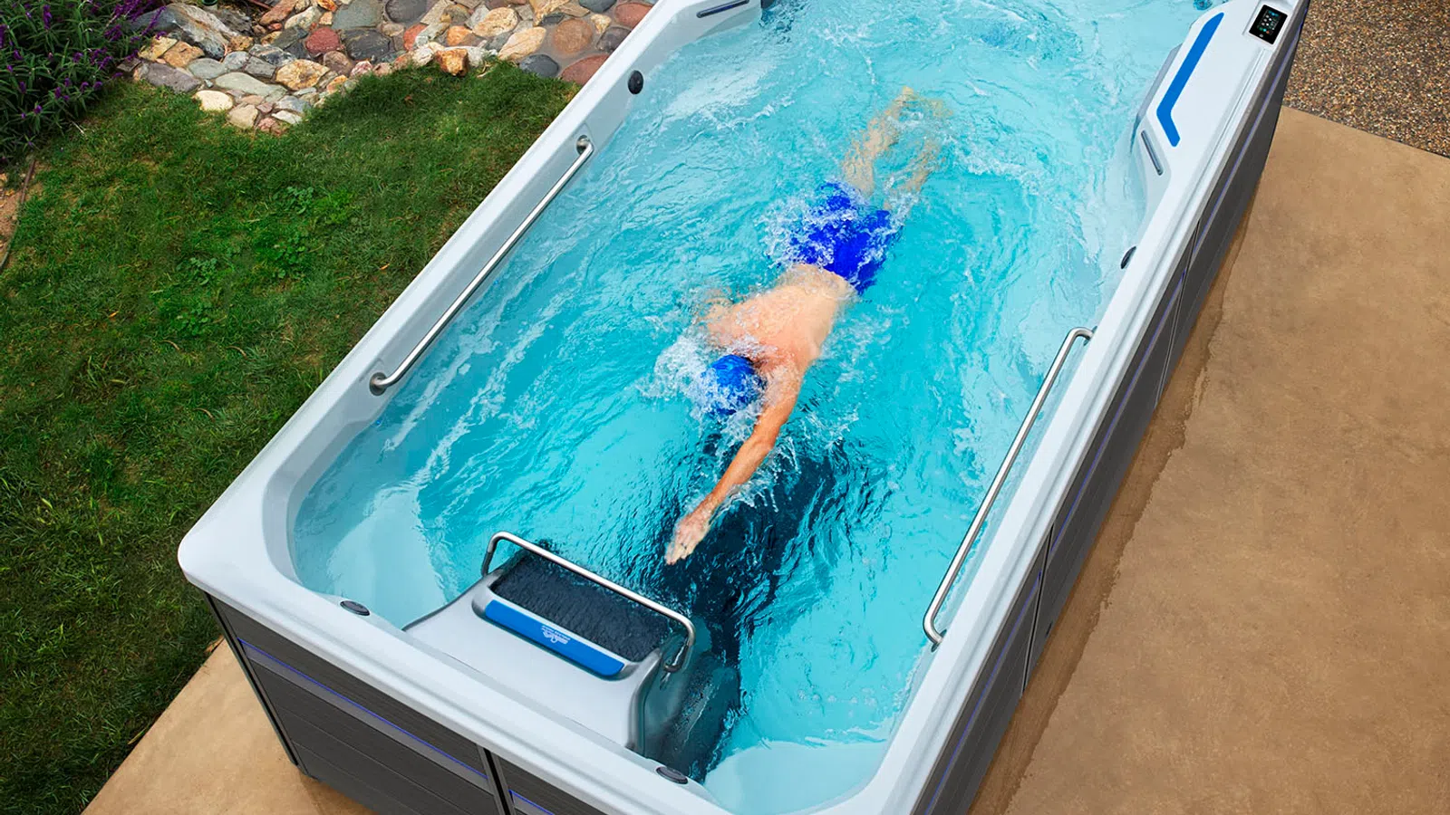 Overhead view of a person swimming in a rectangular outdoor swim spa surrounded by garden and patio areas.