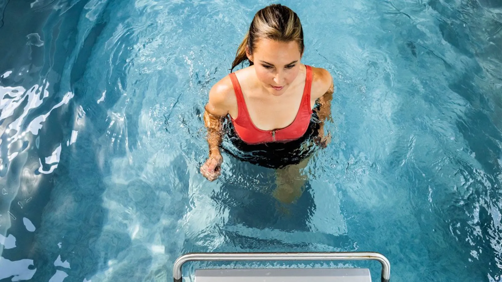 Young woman in red swimsuit standing in clear blue swimming pool near metal ladder steps