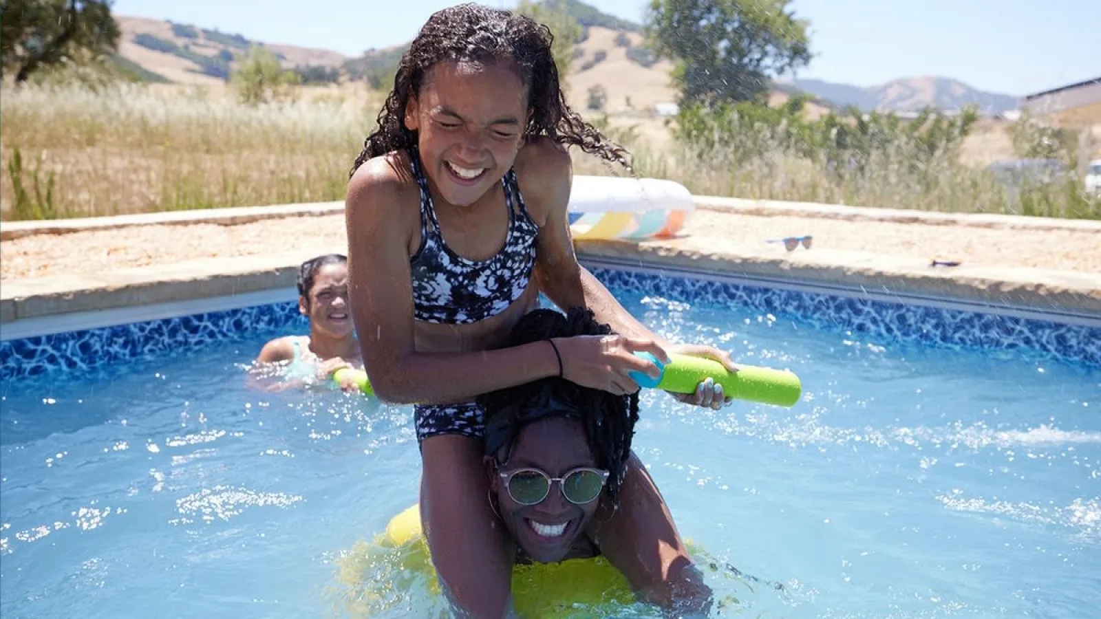 Two girls playing with water guns and smiling in a backyard swimming pool on a sunny day.