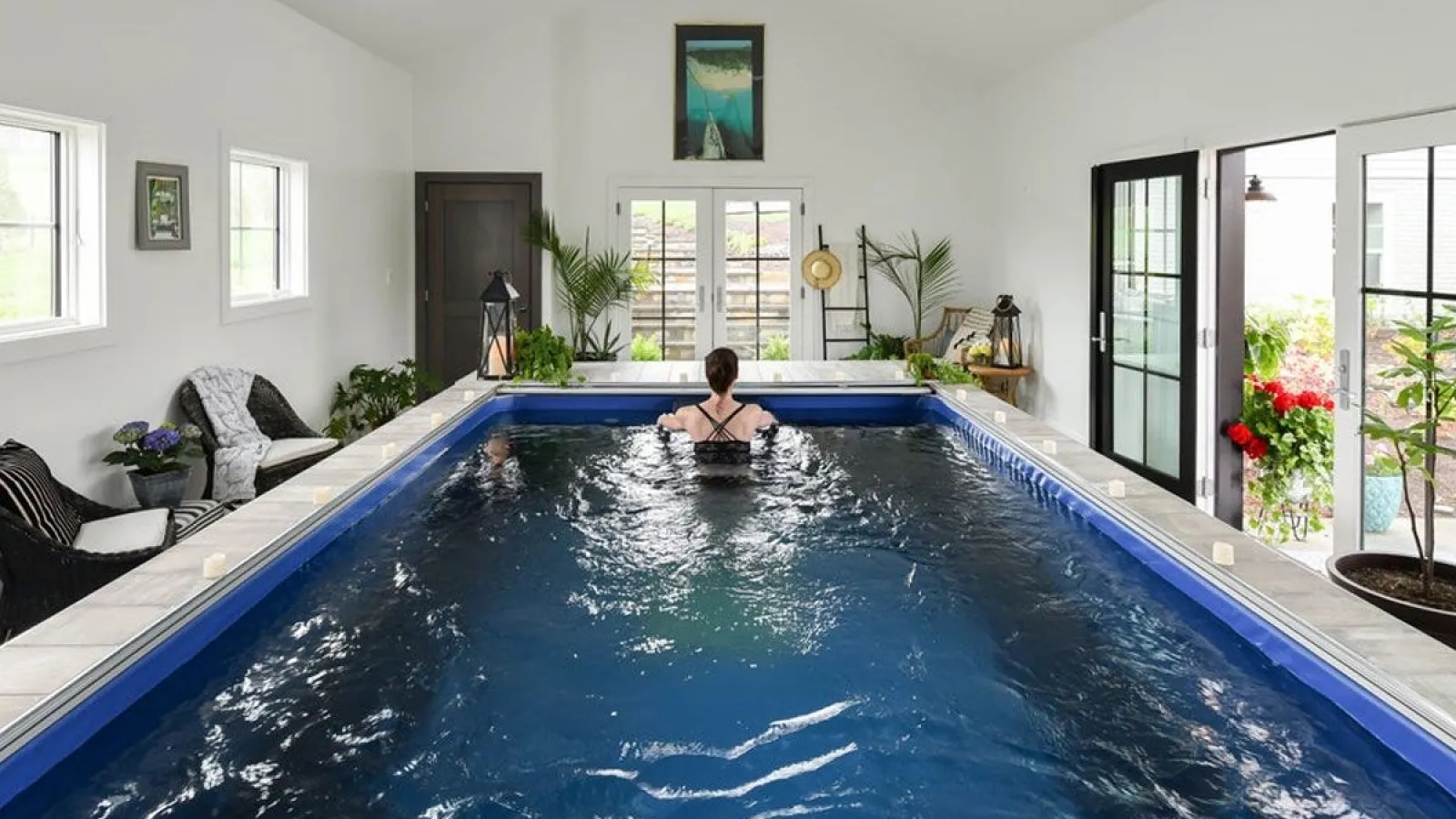 Indoor swimming pool with a woman relaxing in water surrounded by white walls, plants, and cozy seating area.