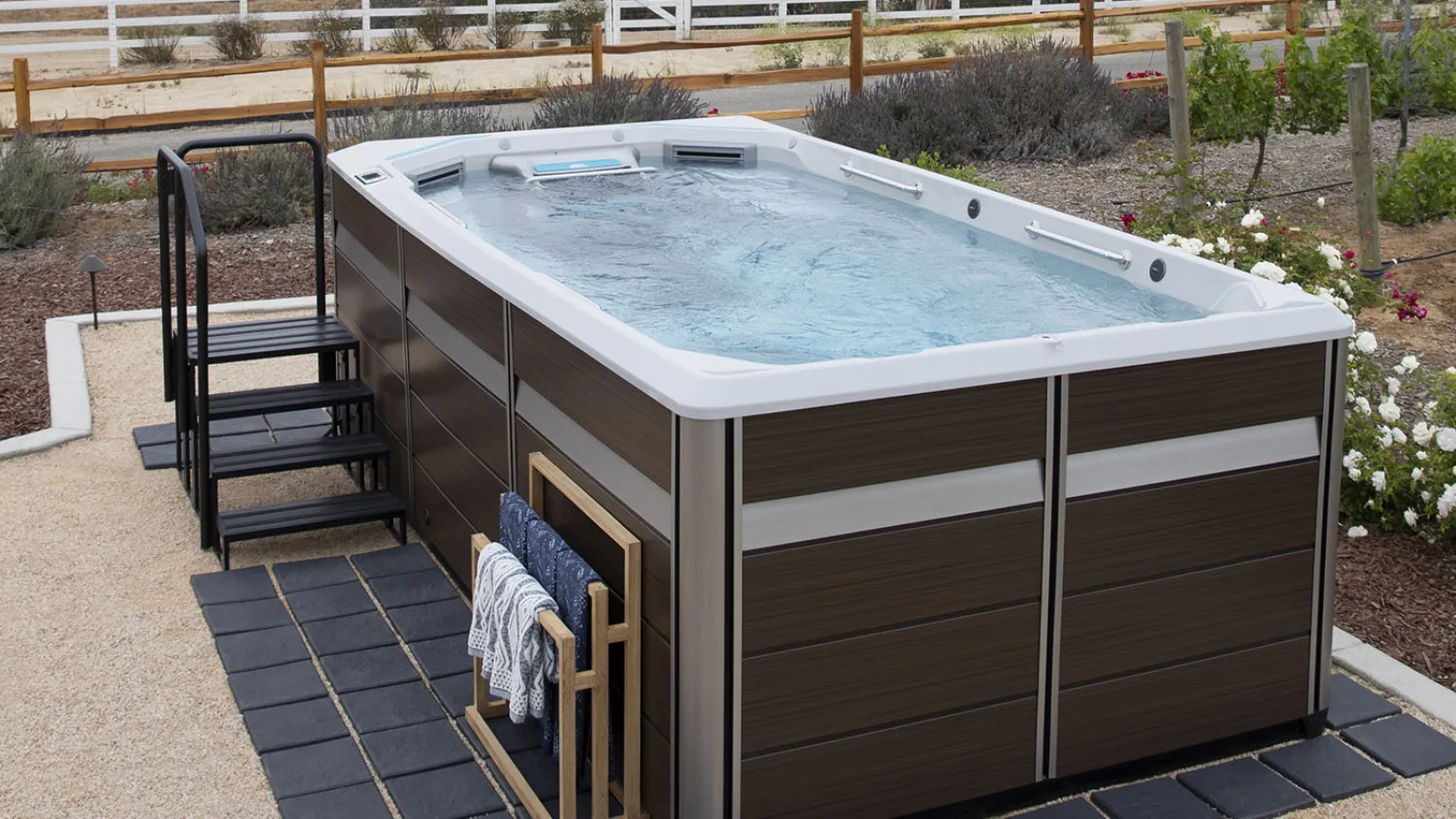 Modern outdoor swim spa with water jets, brown panels, steps, and towel rack in landscaped backyard patio