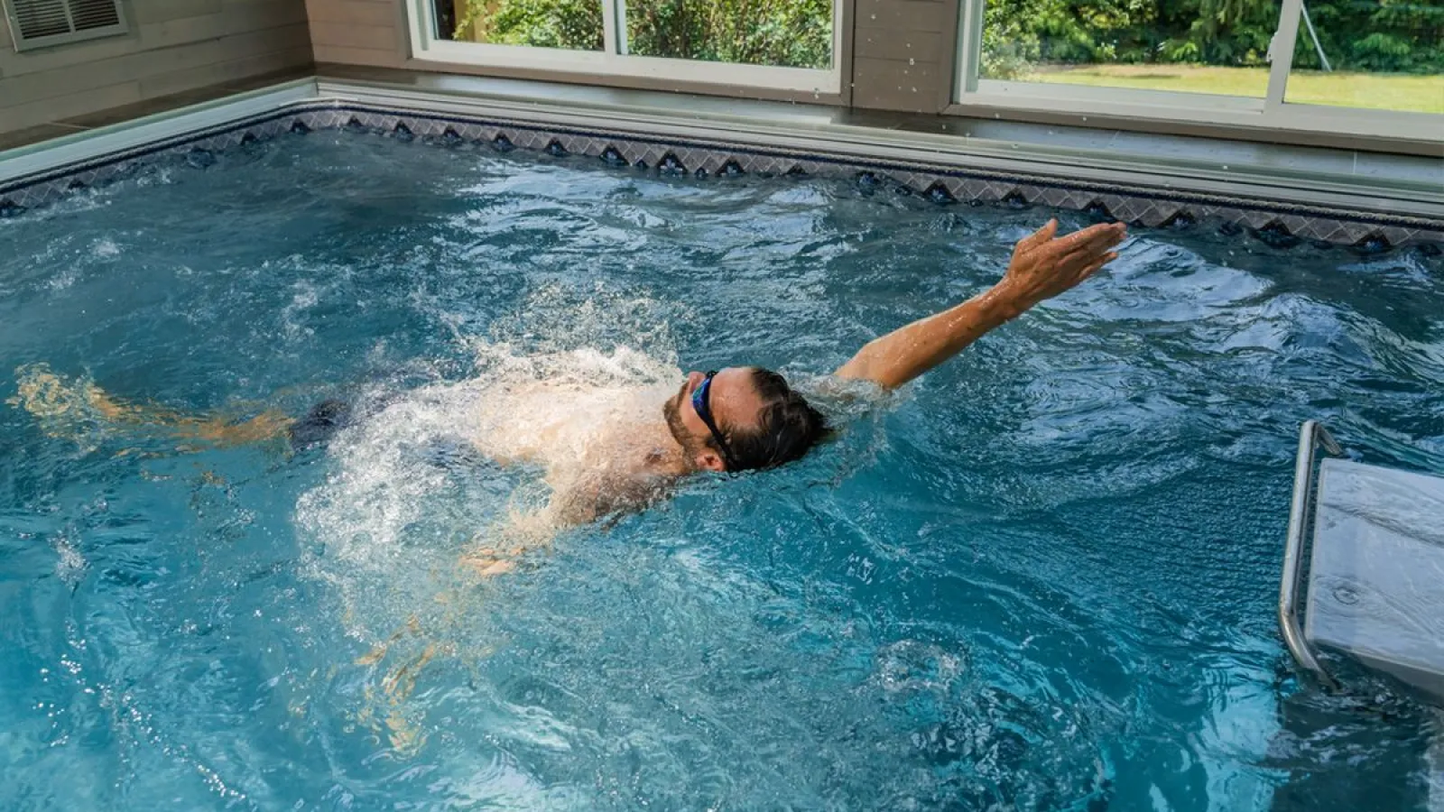 Man wearing goggles swimming backstroke in an indoor pool with sunlight from windows.