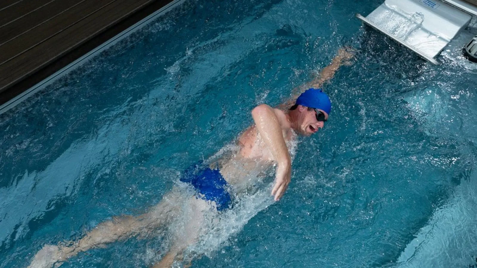 Man swimming freestyle in an indoor pool wearing blue swim cap and goggles, creating splashes in water.