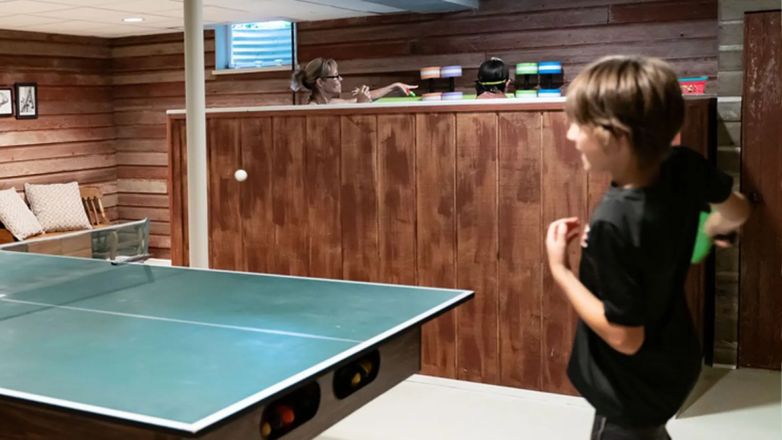 Boy playing table tennis in a wood-paneled basement with two people watching over a wooden divider.