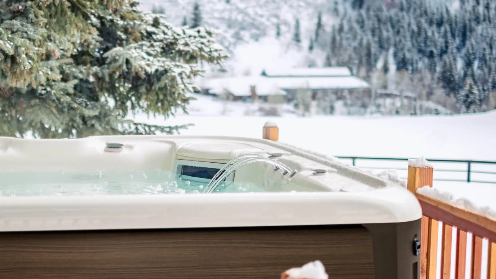 Outdoor hot tub on wooden deck with snow-covered trees and mountains in winter landscape background