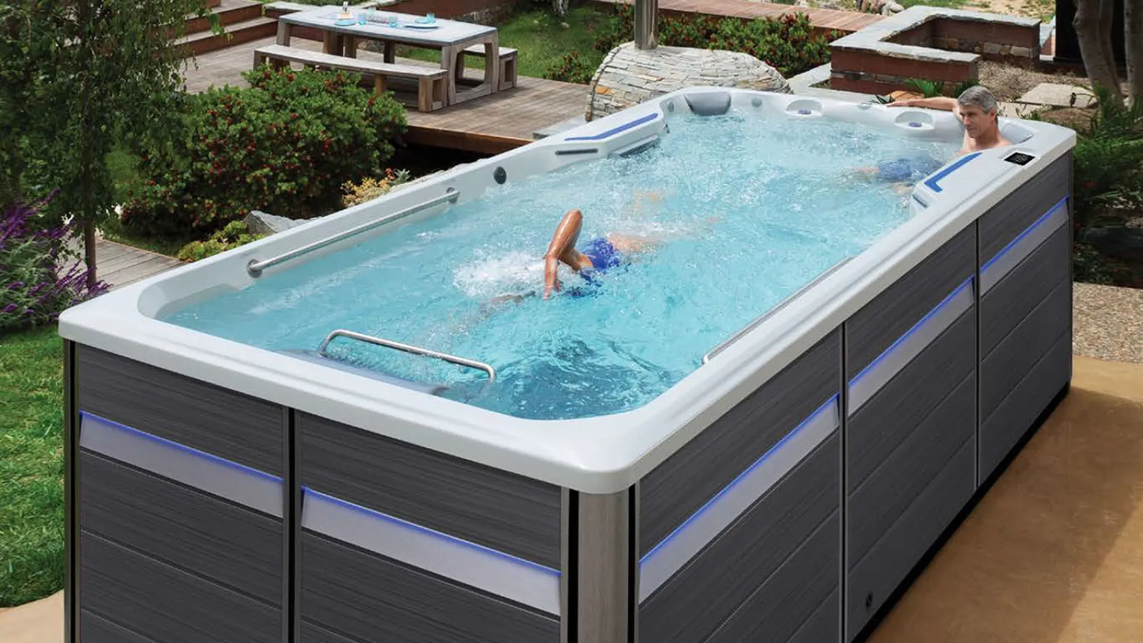 Modern outdoor swim spa with a person swimming, surrounded by greenery and patio seating area.