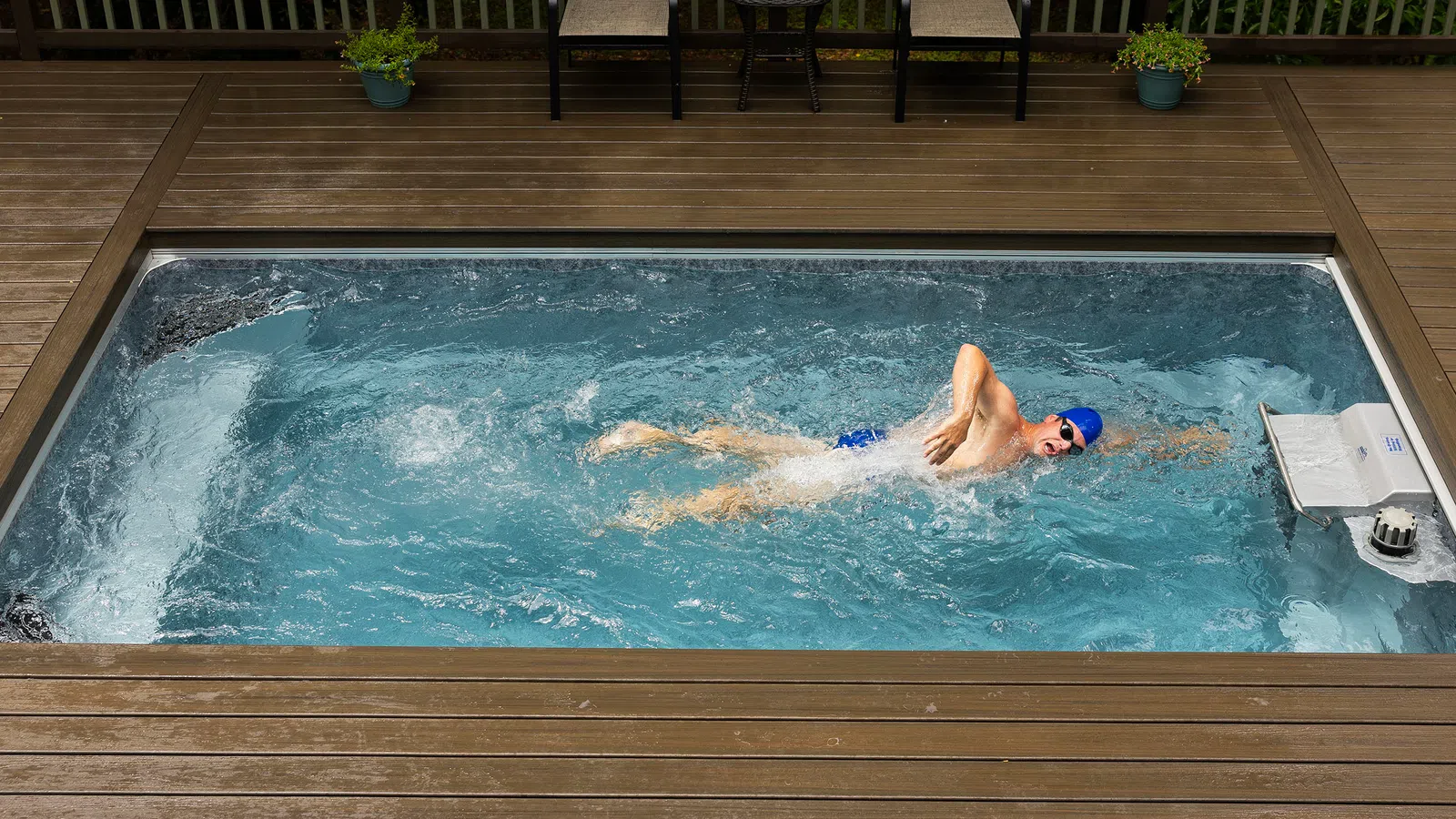 Swimmer in blue cap swimming in a rectangular indoor pool surrounded by wooden deck and seating area.