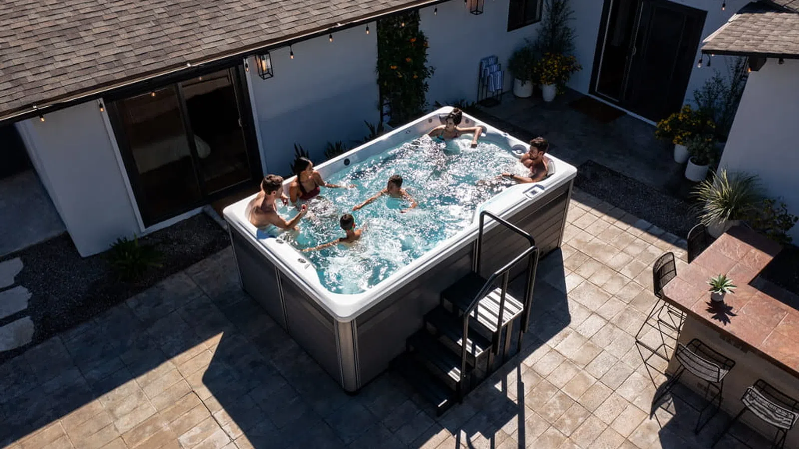 Family of six enjoying a backyard hot tub on a sunny tiled patio beside a house with potted plants.