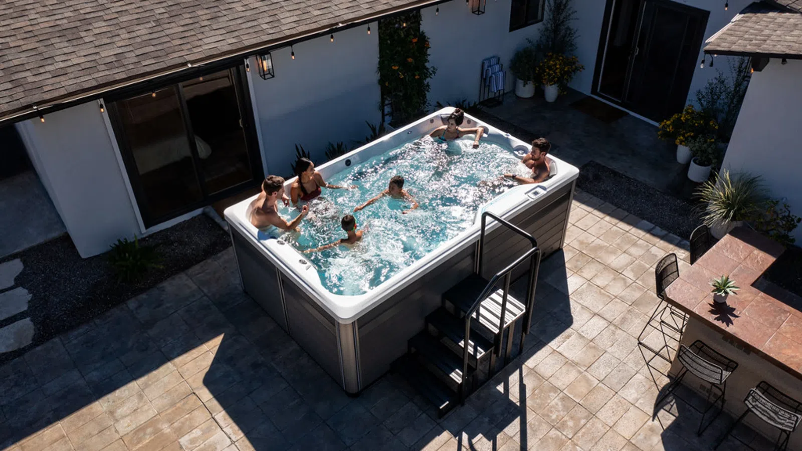 Overhead view of six people enjoying a large hot tub in a sunny courtyard patio beside a house.