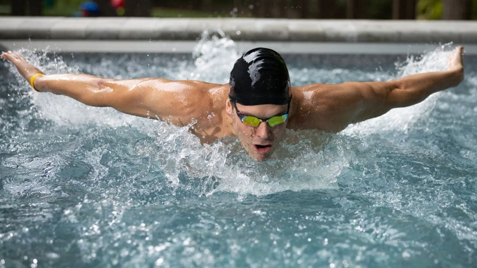 Male swimmer in black cap and goggles performing butterfly stroke in outdoor pool water splashing