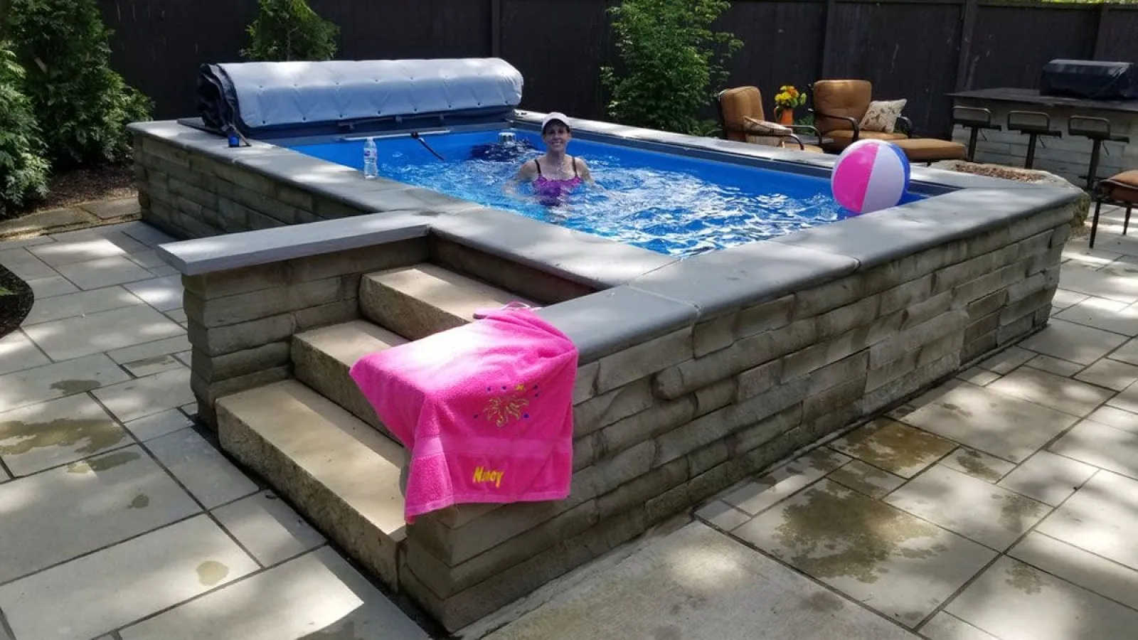 Woman swimming in an above-ground stone pool with pink towel and beach ball in a sunny backyard patio.