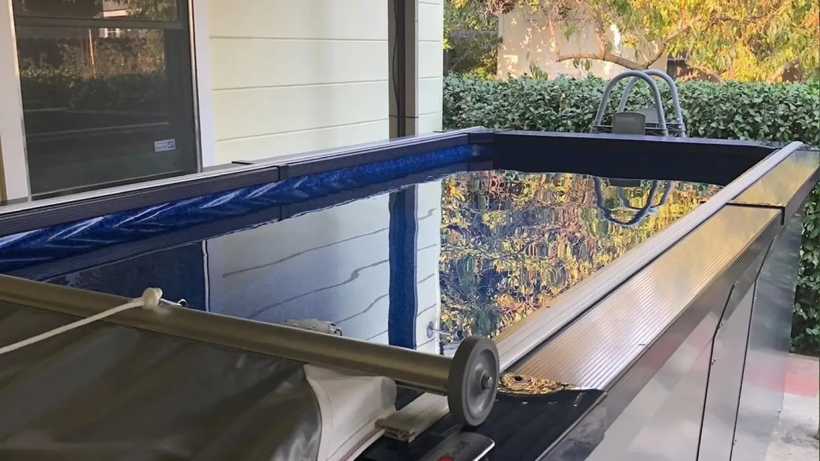 Above-ground rectangular pool with a blue water cover partially rolled, reflecting nearby foliage and house exterior.