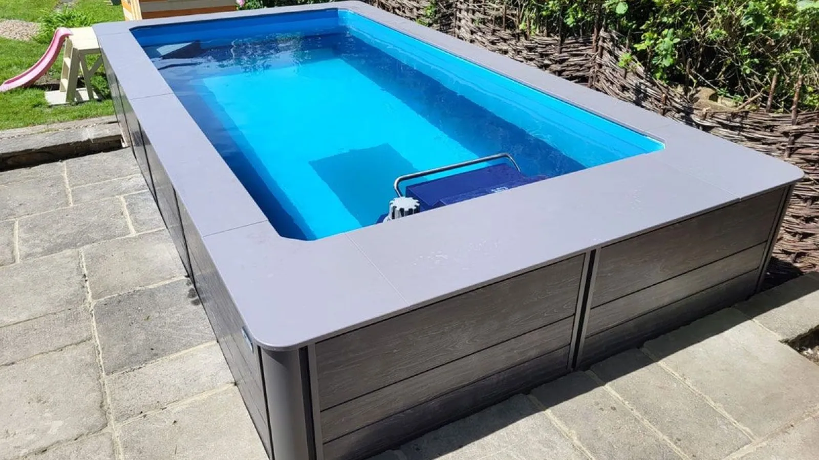 Rectangular above-ground swimming pool with clear blue water in a backyard with stone patio and wicker fence.