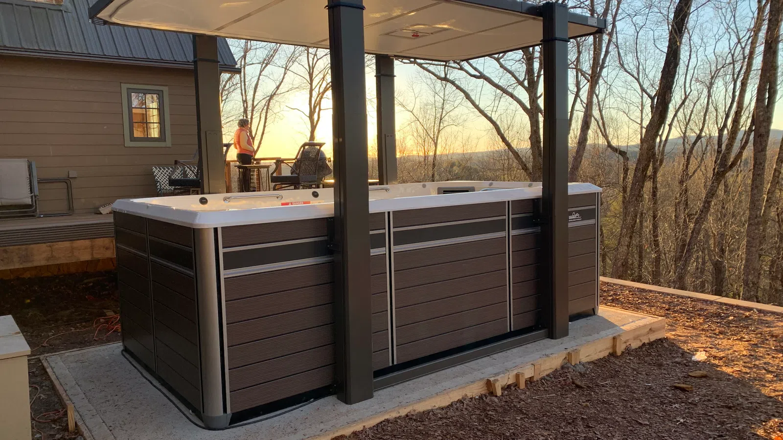 Outdoor hot tub with a canopy on a concrete slab and a house in the background at sunset.
