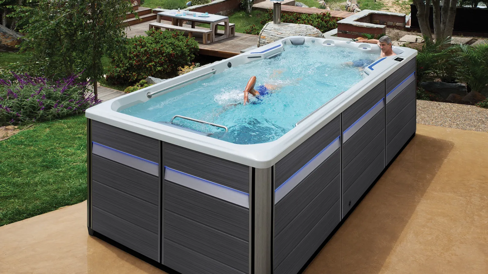 Outdoor swim spa with one person swimming and another relaxing, surrounded by garden and patio furniture.