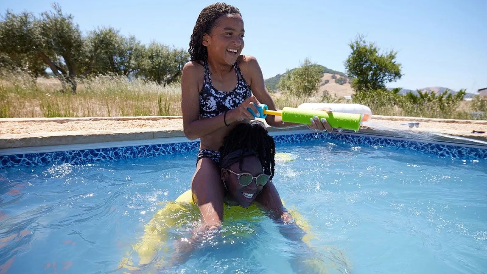 Two children playing joyfully with a water gun in a backyard swimming pool on a sunny day