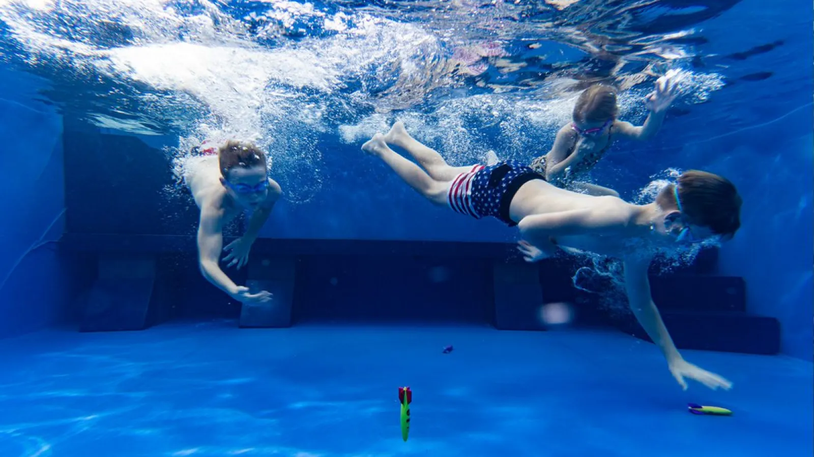 Three children swimming underwater in a pool wearing goggles and chasing colorful diving toys.