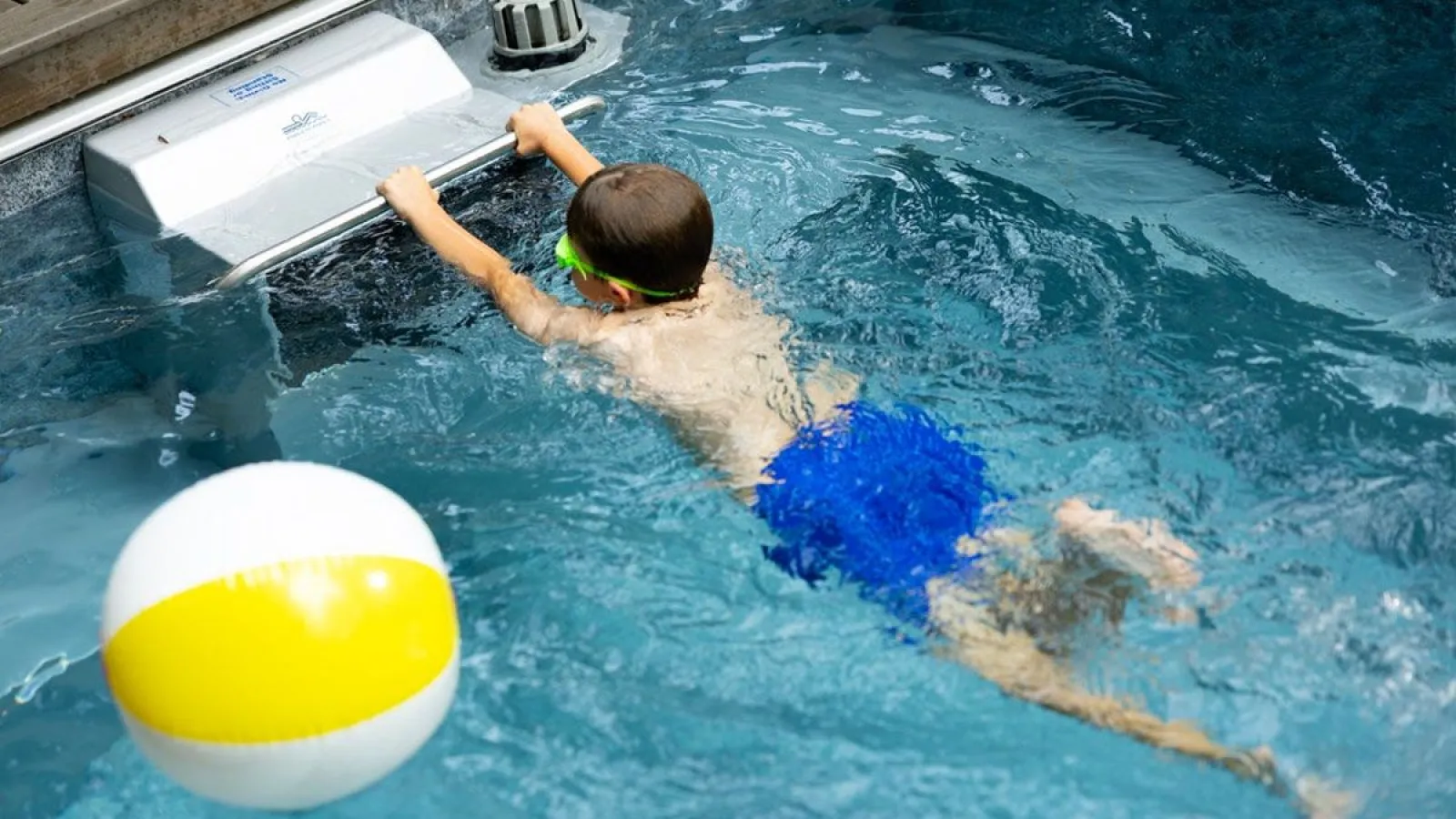 Boy in blue swim trunks using a pool swim jet while a yellow and white beach ball floats nearby.