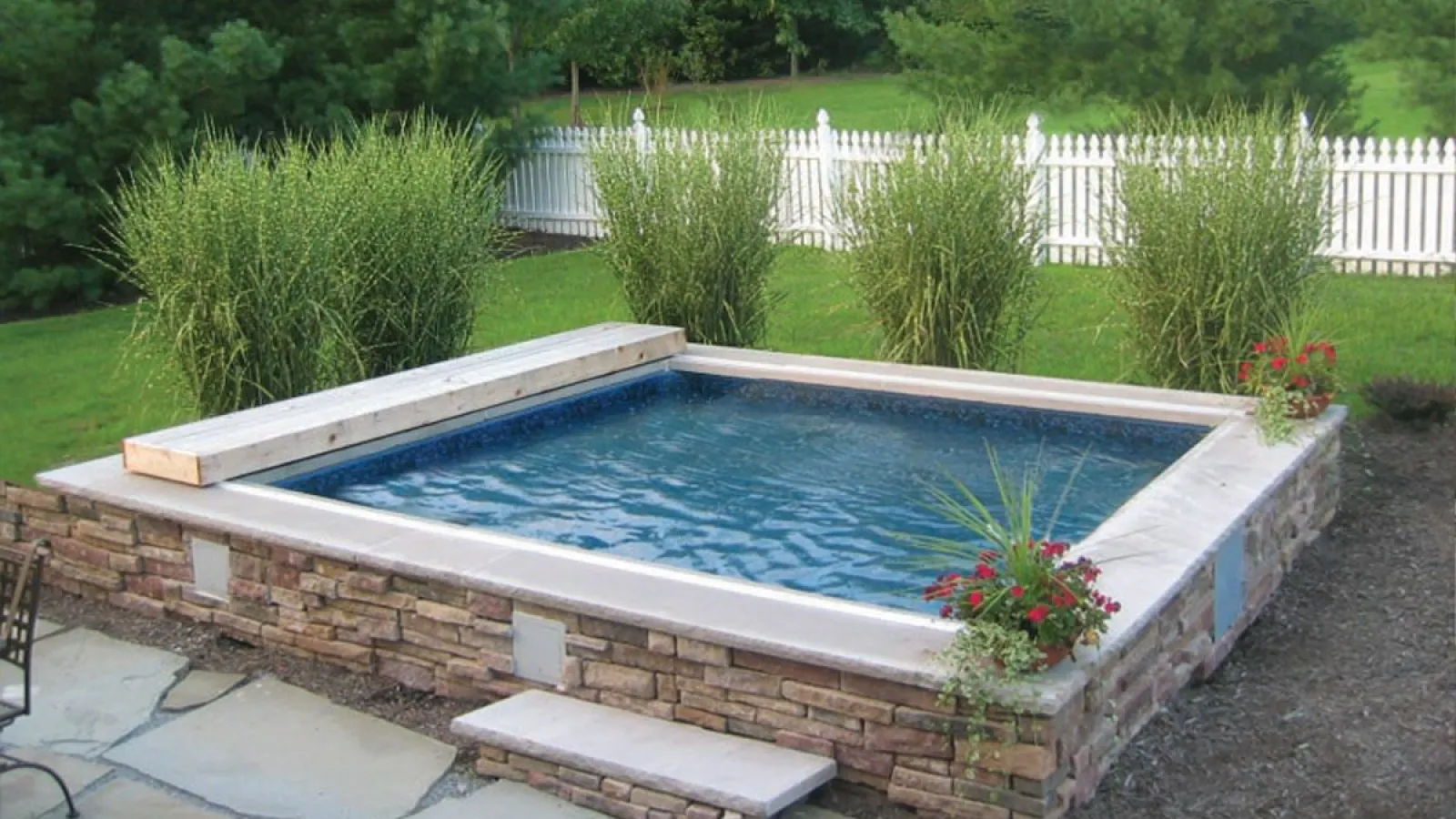 Small rectangular stone hot tub with clear water surrounded by tall grass and white picket fence