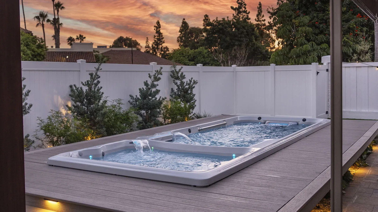 Modern backyard swim spa with water features and ambient lighting under a colorful sunset sky.