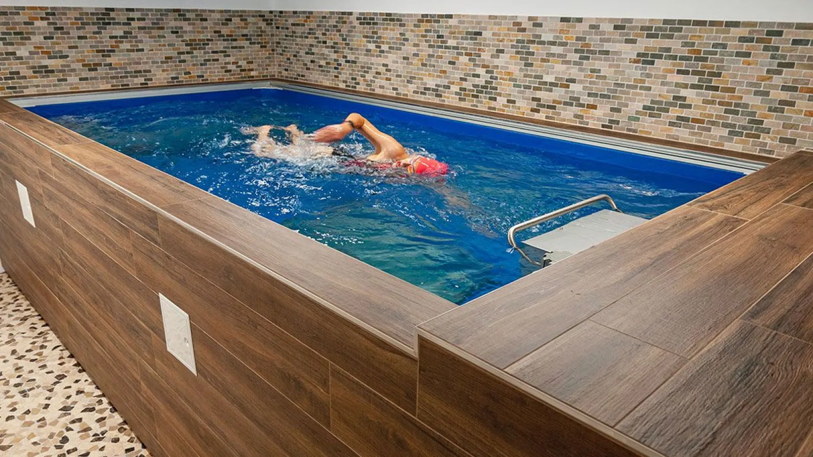 Indoor swimming pool with wood-paneled walls and tile backsplash featuring a person swimming freestyle with a red cap.