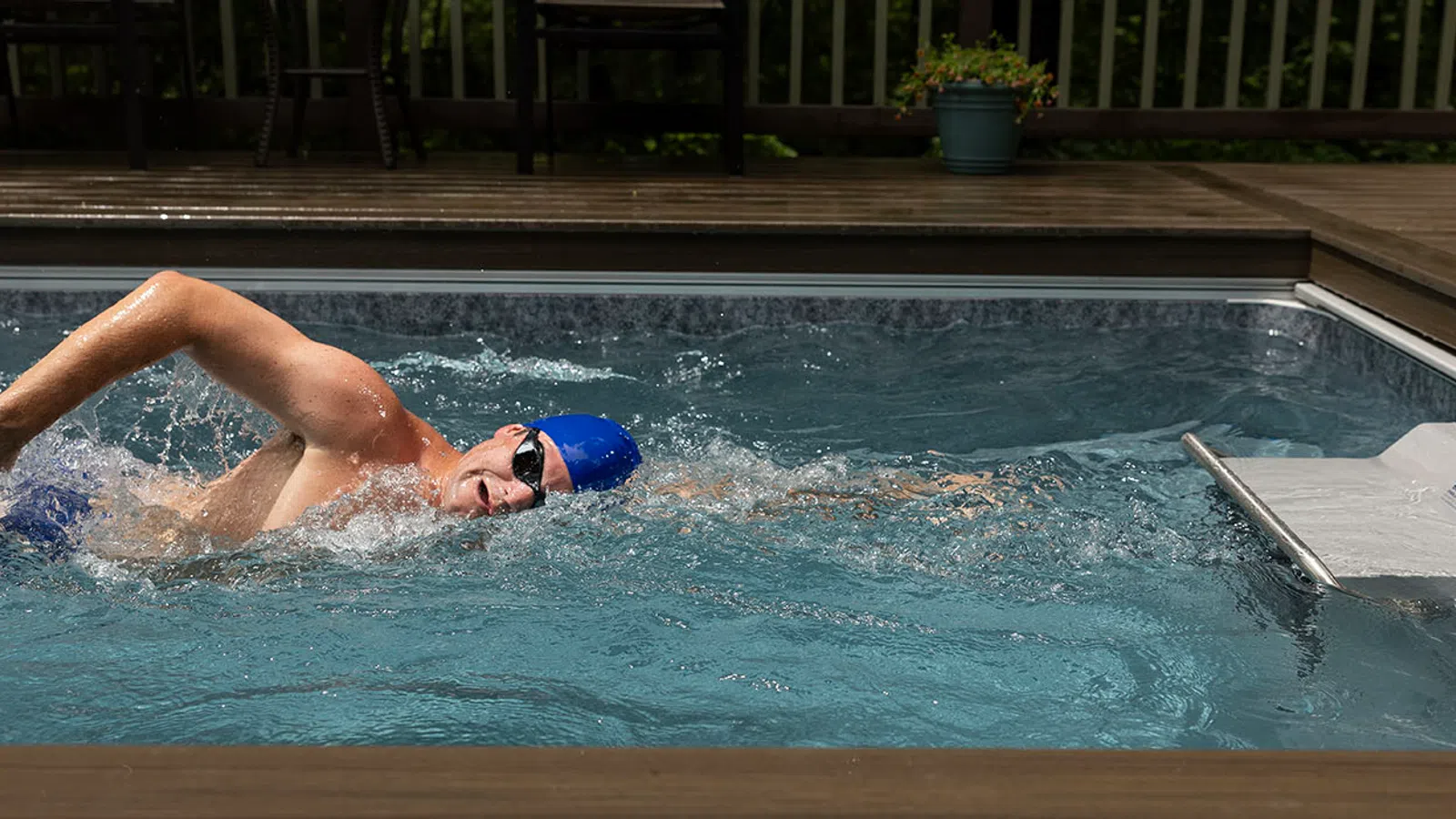 Man swimming freestyle in a backyard pool with wooden deck and blue swim cap and goggles.
