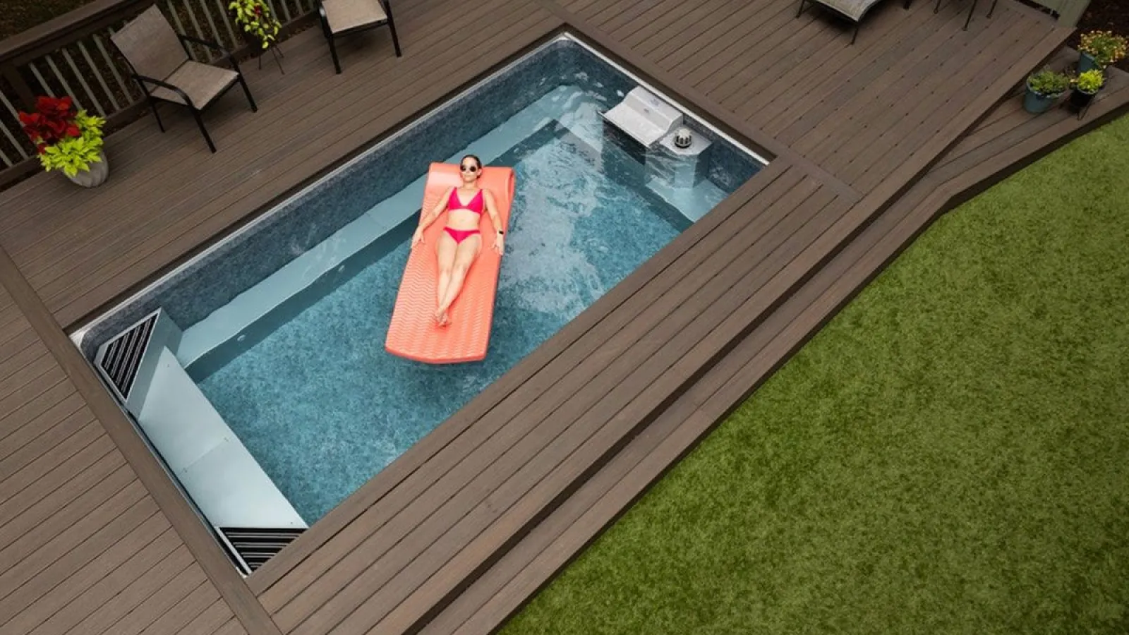 A woman in a red swimsuit relaxes on an orange float in a small, modern pool surrounded by wooden decking and green grass.