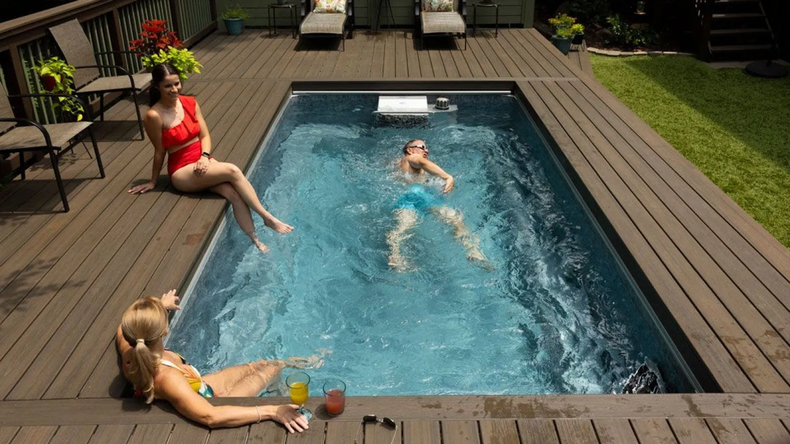 People enjoying a backyard swim spa with wooden deck, two women sitting and a man swimming inside the pool.