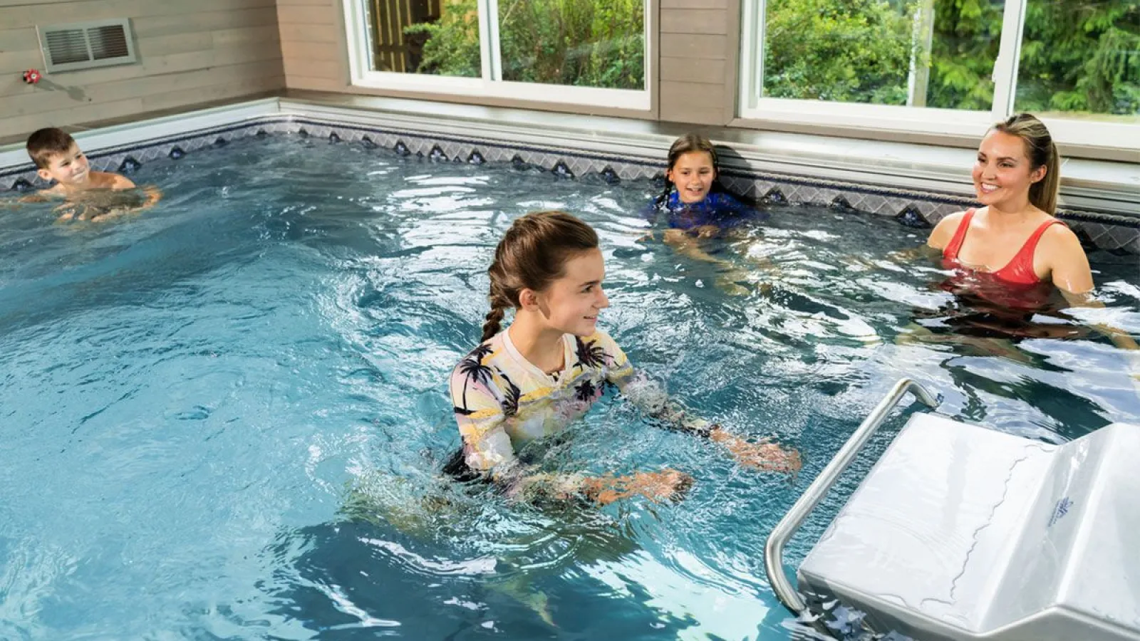 Children and a woman enjoying time together in an indoor pool with large windows and greenery outside.