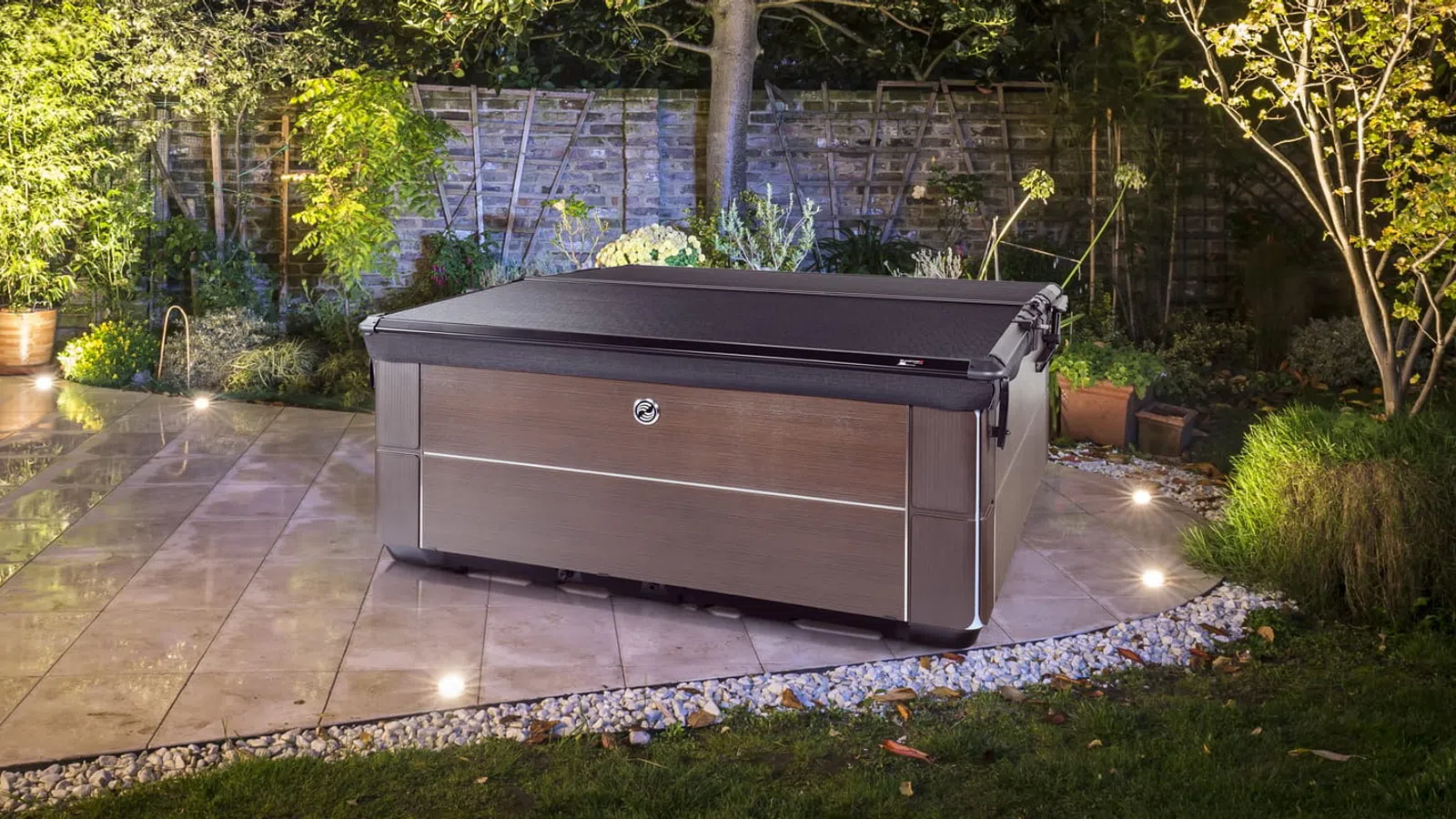 Modern outdoor hot tub with closed cover on tiled patio surrounded by garden and ambient lighting at night