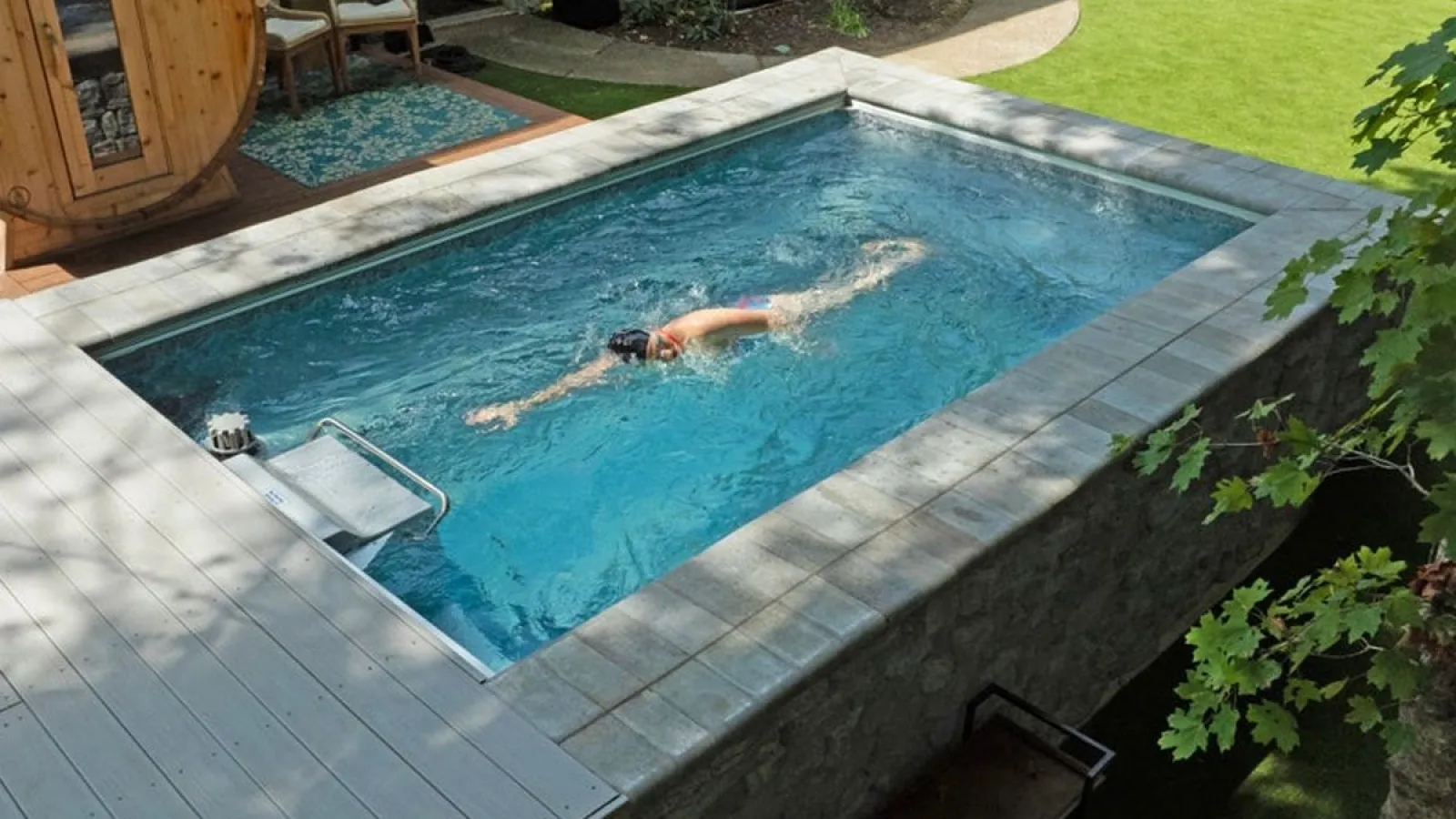 Person swimming in a modern rectangular backyard pool with stone and wood decking near green lawn.