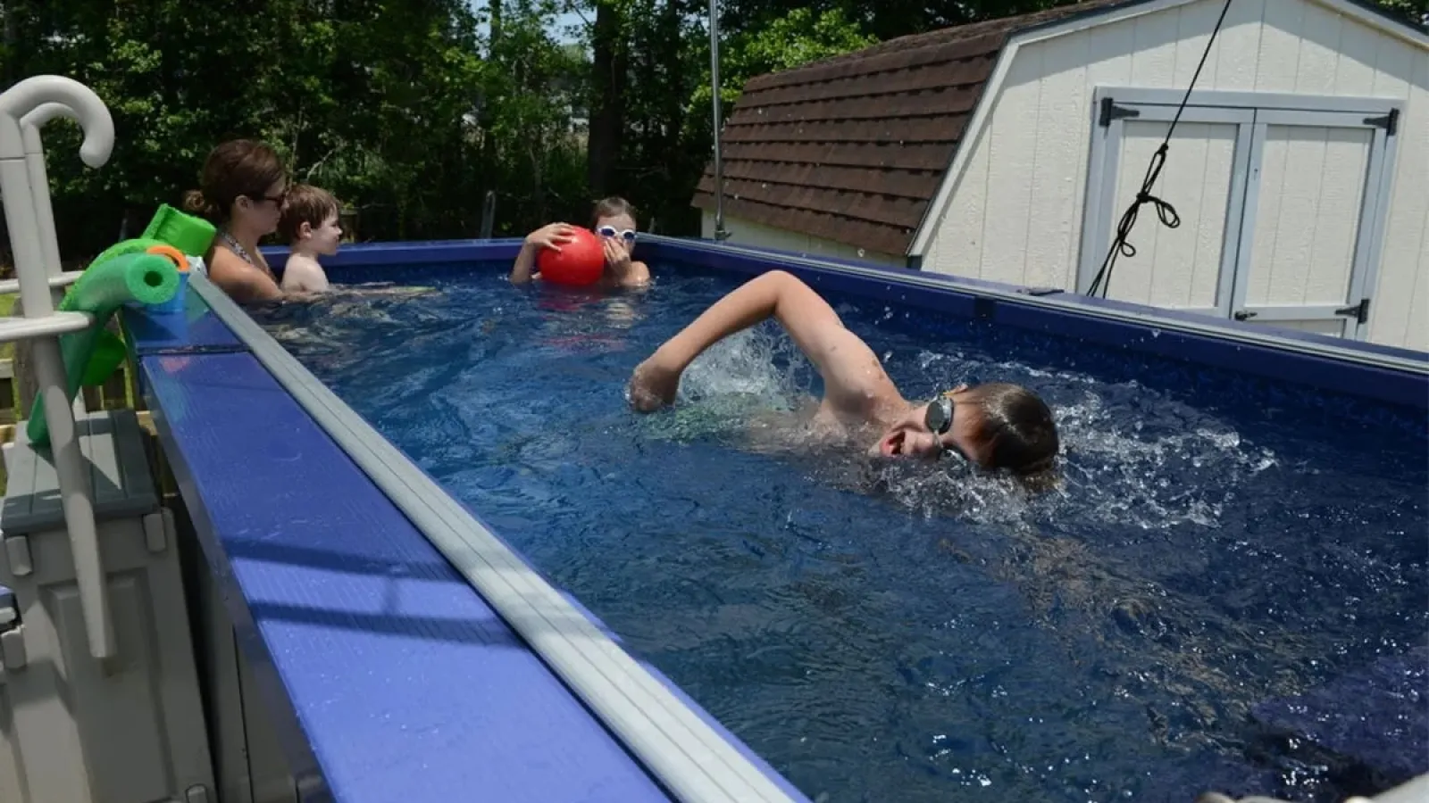Children swimming and playing in a blue above-ground pool near a white shed on a sunny day.