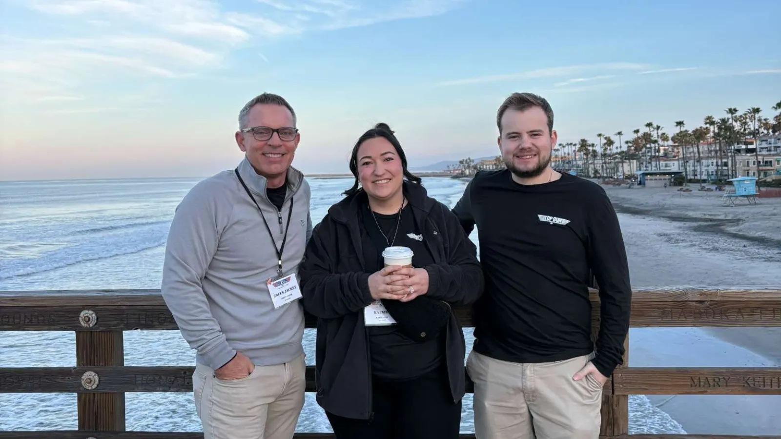 Three people standing on a wooden pier smiling with ocean waves and palm trees in the background under a clear sky.