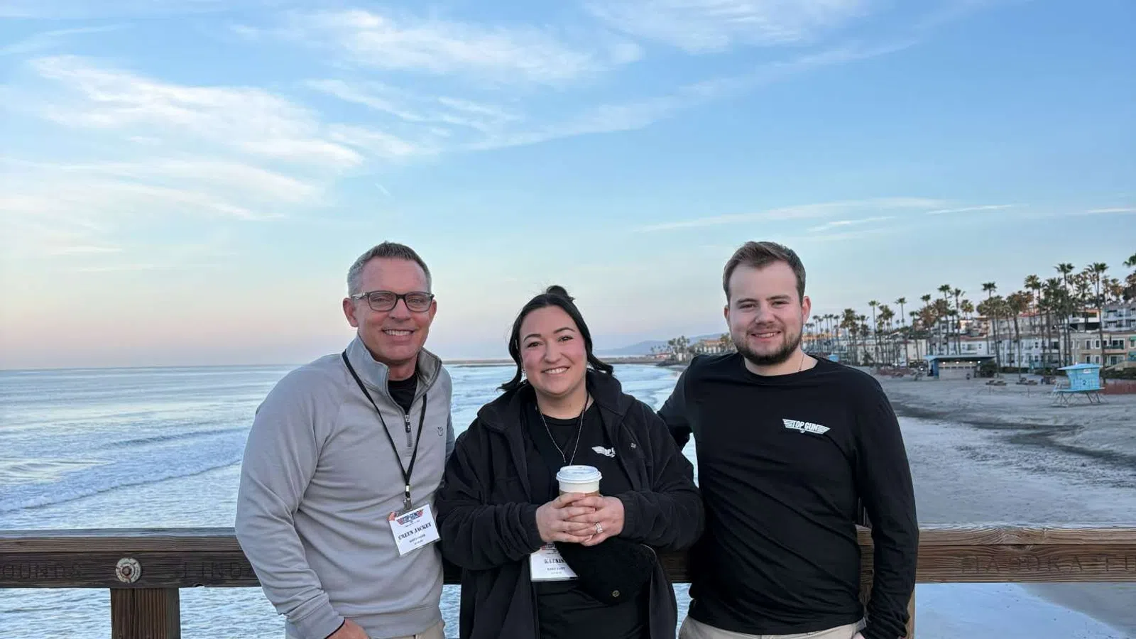 Three people standing on a wooden pier smiling with ocean waves and palm trees in the background under a clear sky.