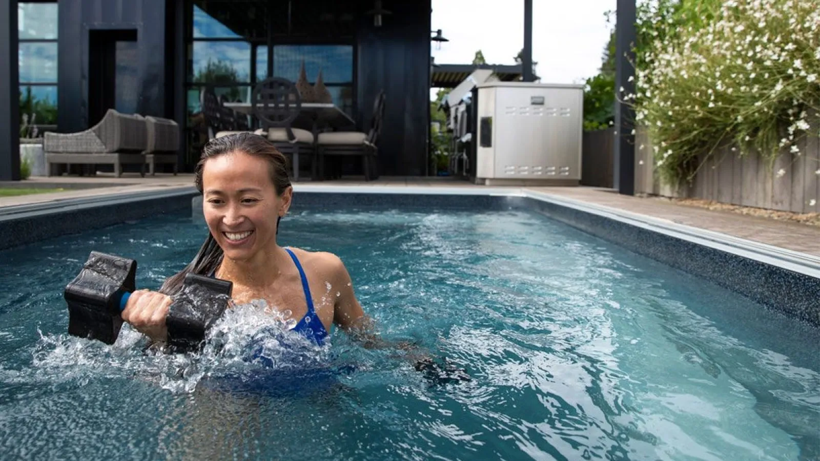 Woman in blue swimsuit exercising with dumbbell in a backyard pool next to outdoor kitchen and seating area.