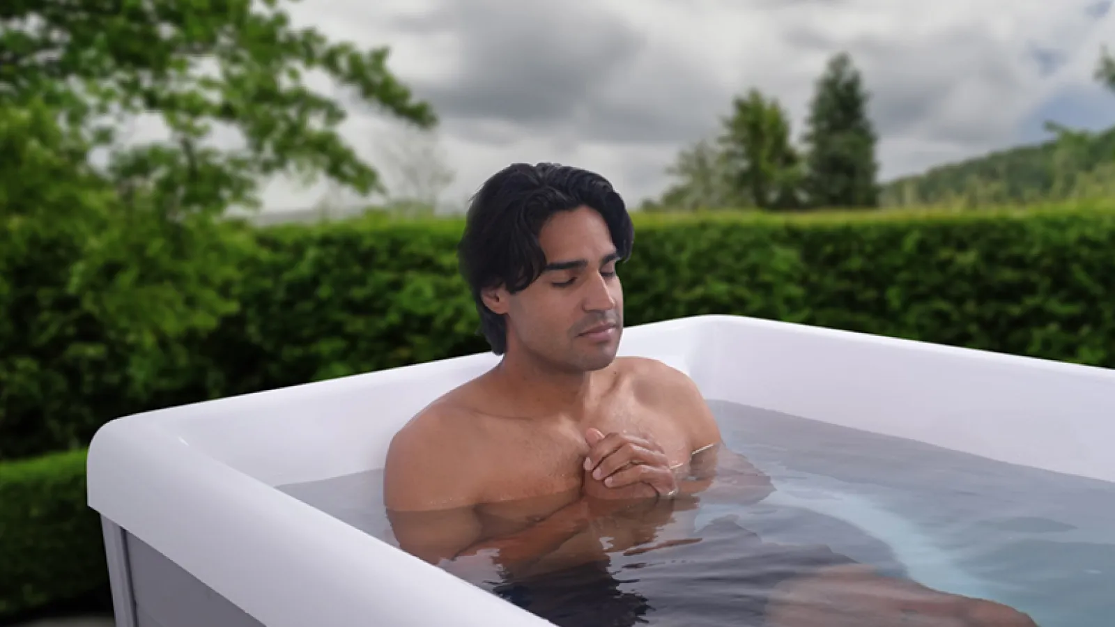 Man relaxing with eyes closed in a modern outdoor hot tub surrounded by greenery on a cloudy day