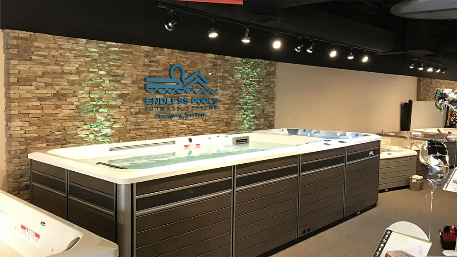 Modern Endless Pools swim spa with water jets displayed inside showroom with stone wall and ceiling lights