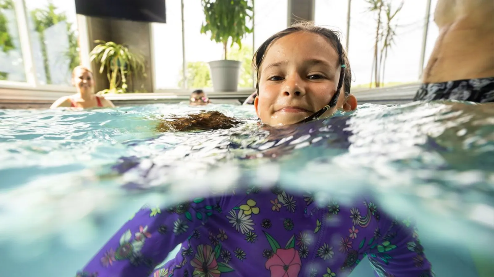 Smiling girl in floral swimwear partially submerged in indoor pool with other swimmers and plants in background