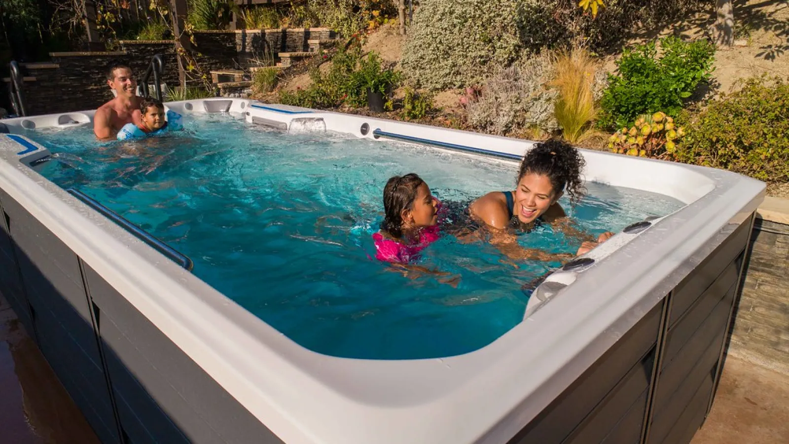 Family swimming and playing in a backyard swim spa with clear blue water on a sunny day.