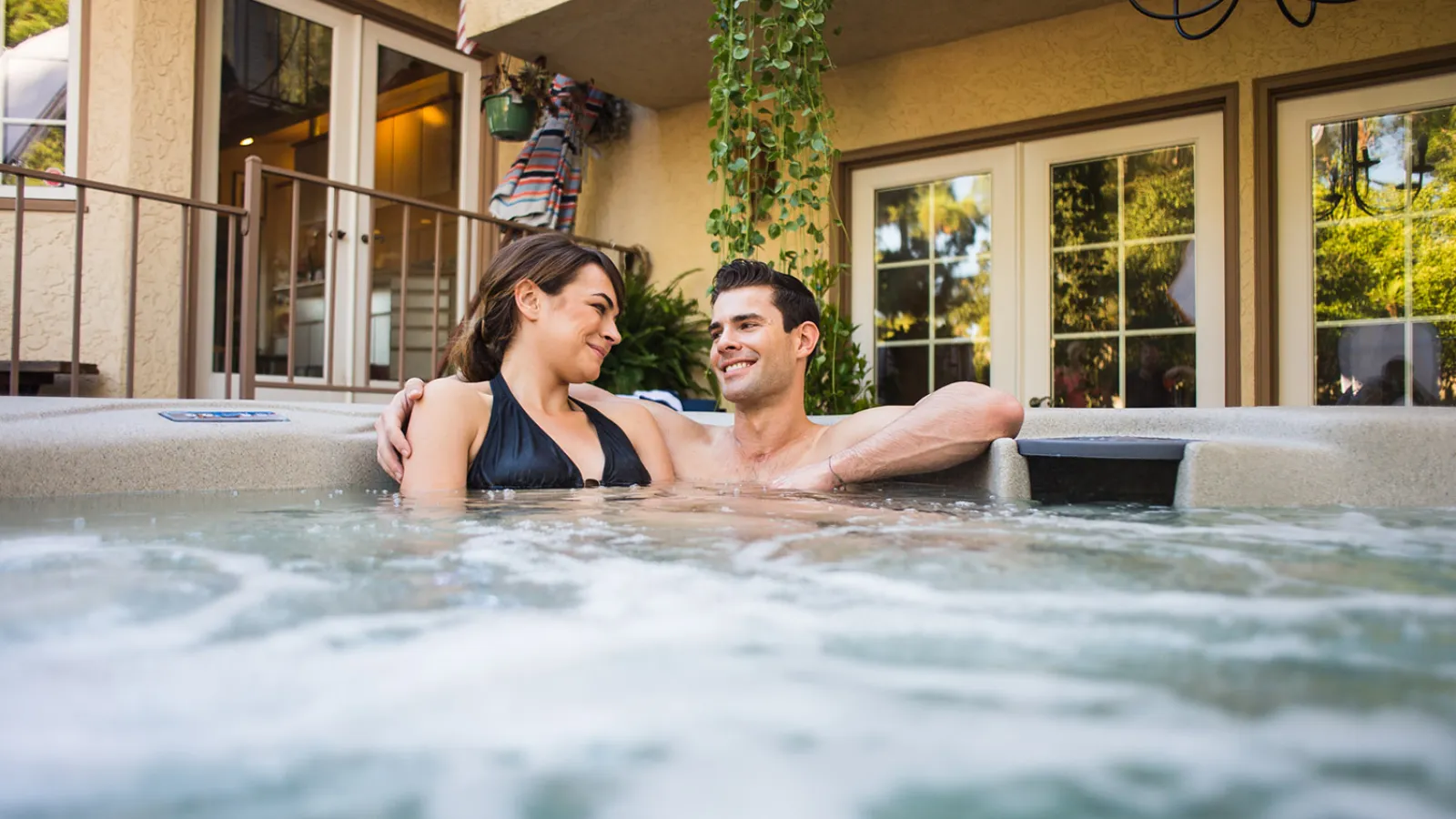Couple enjoying a relaxing moment in a hot tub surrounded by lush greenery and sunlight.
