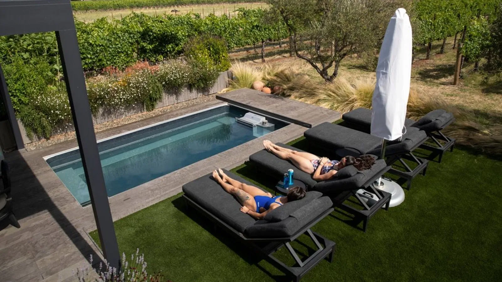 Two people relaxing on black lounge chairs beside a small rectangular pool with greenery and vineyard backdrop.