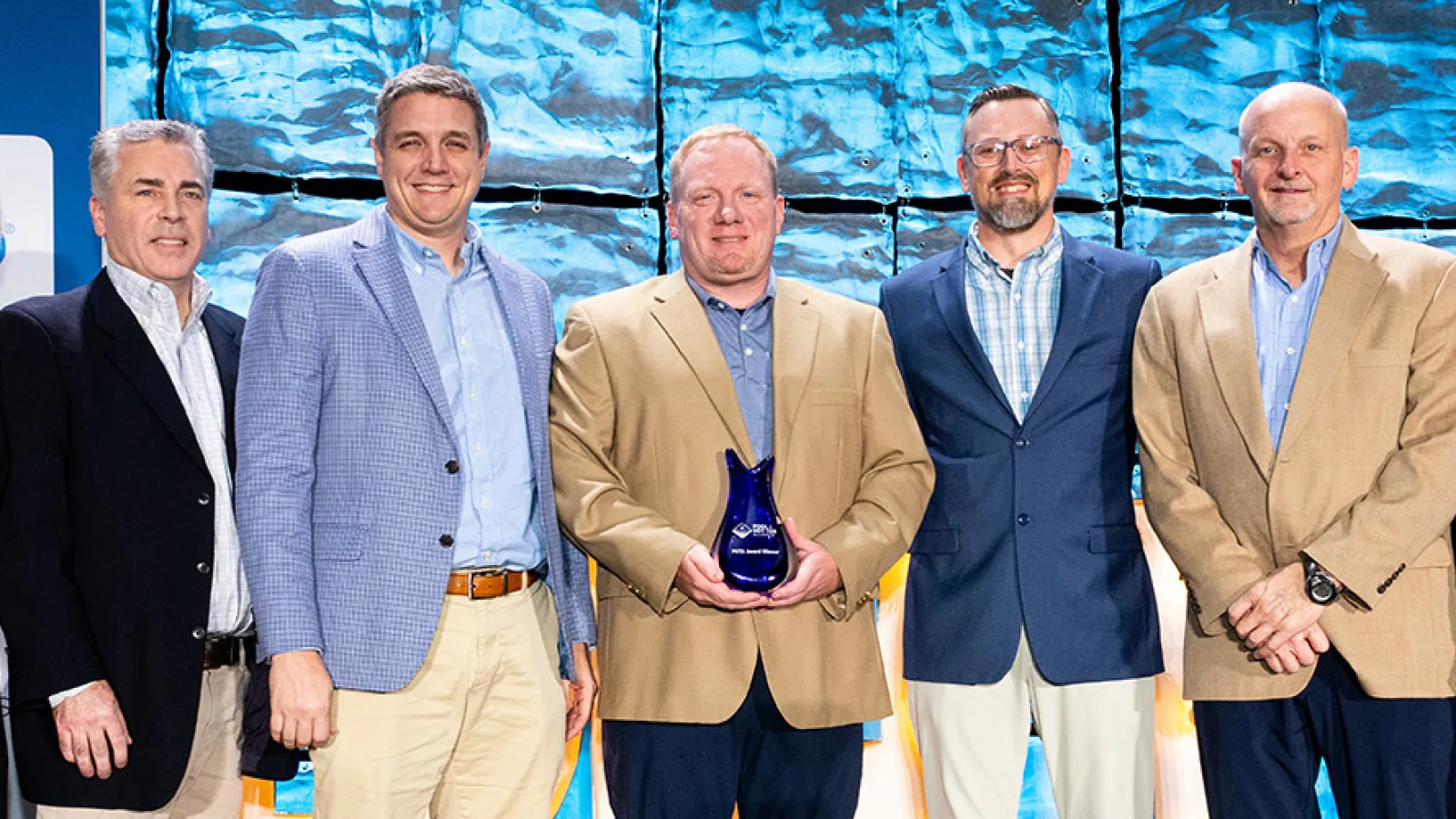 Group of seven professionally dressed people standing on stage with man in center holding blue award trophy