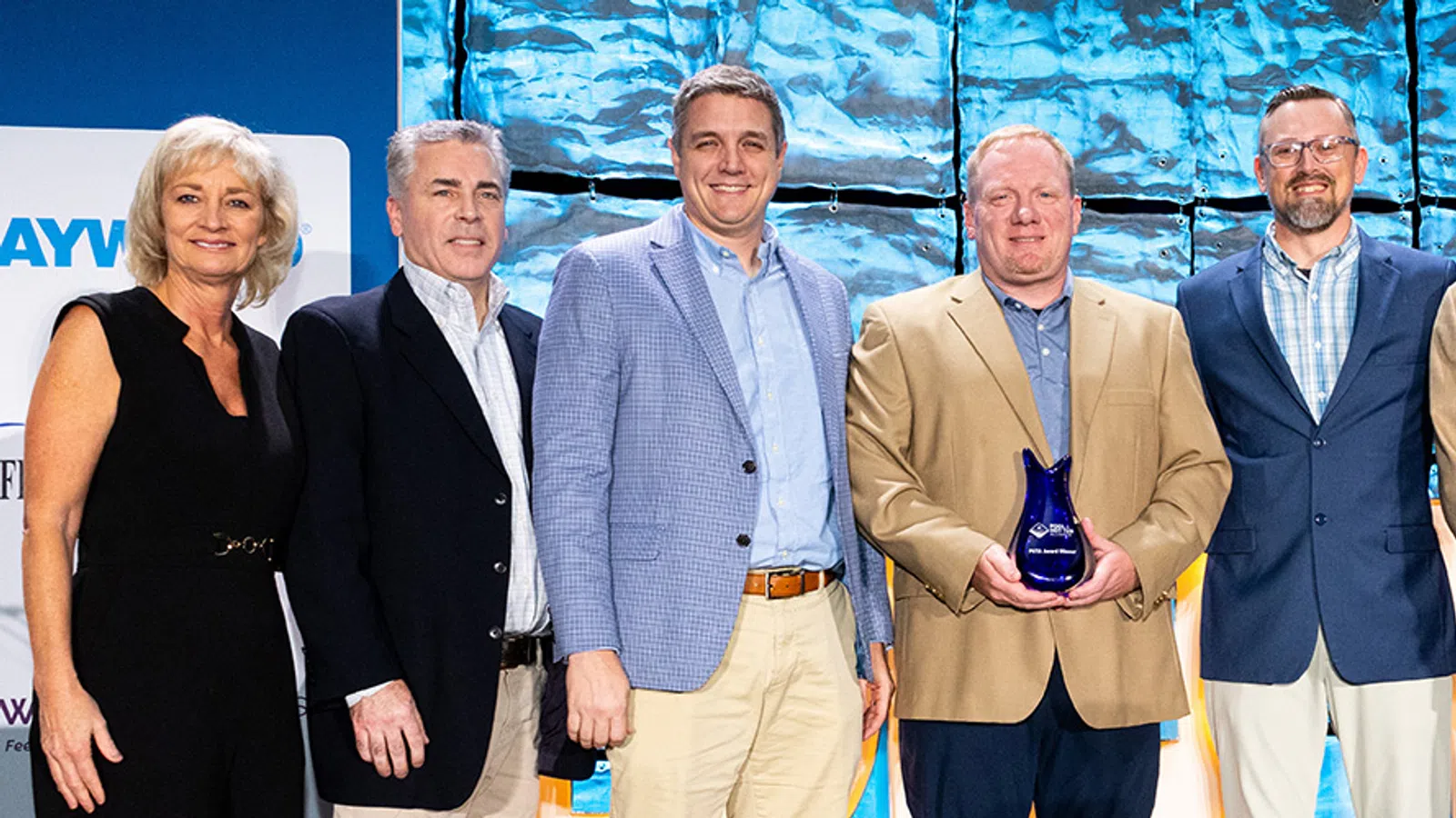Group of seven professionally dressed people standing on stage with man in center holding blue award trophy