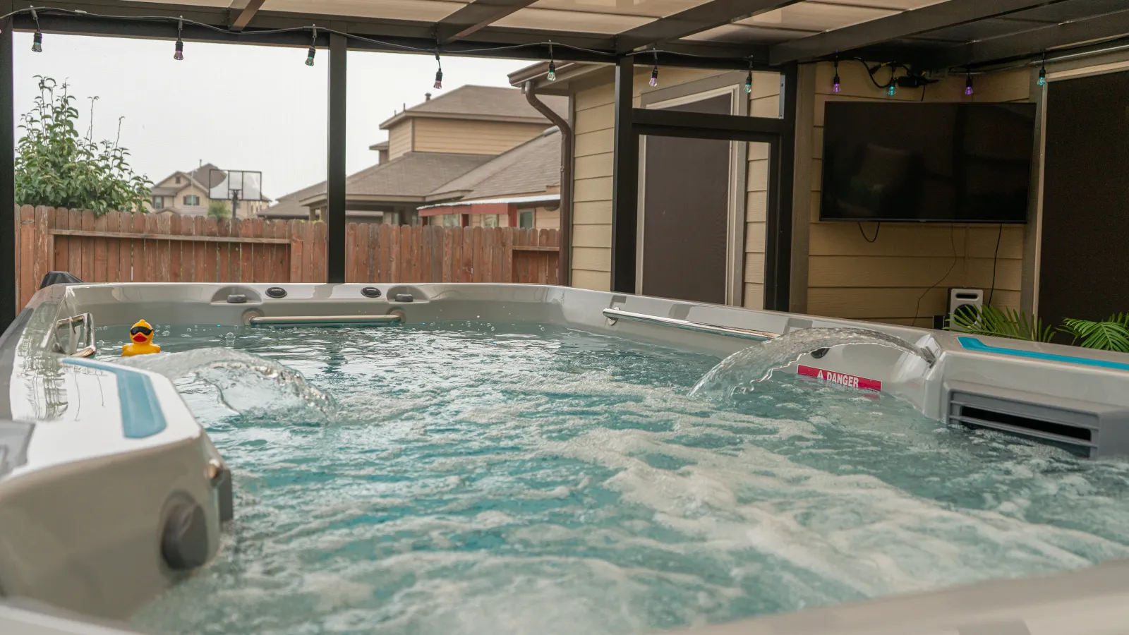 Outdoor covered swim spa with bubbling water and a rubber duck under a patio roof in a backyard.