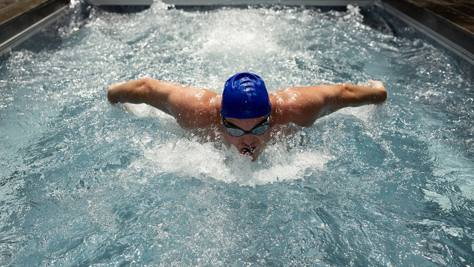 Swimmer in blue cap and goggles performing butterfly stroke in a pool with splashing water.
