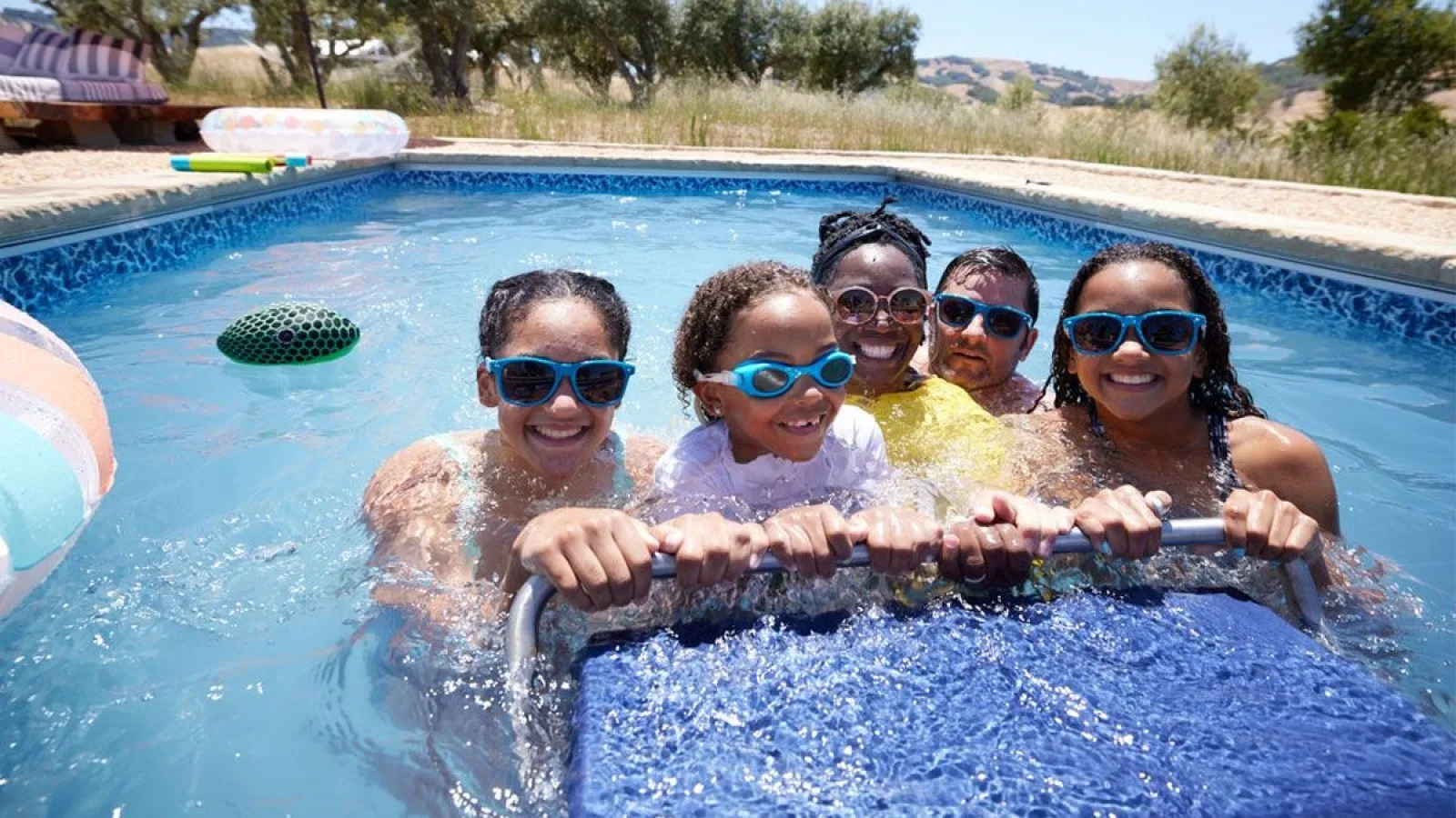 Five people wearing goggles and sunglasses smiling while holding onto a pool float in a sunny backyard pool.