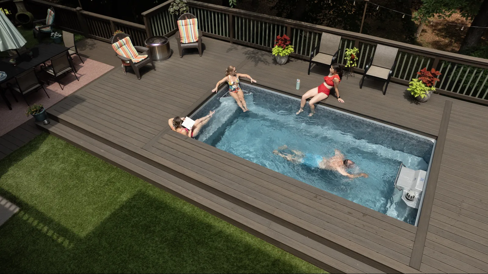 Four people enjoying a small rectangular pool surrounded by a wooden deck with chairs and plants.